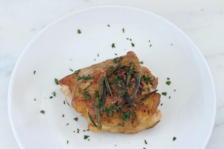 A single bone-in, skin-on chicken thigh served on a white plate with rosemary, pan juices, and chopped parsley.