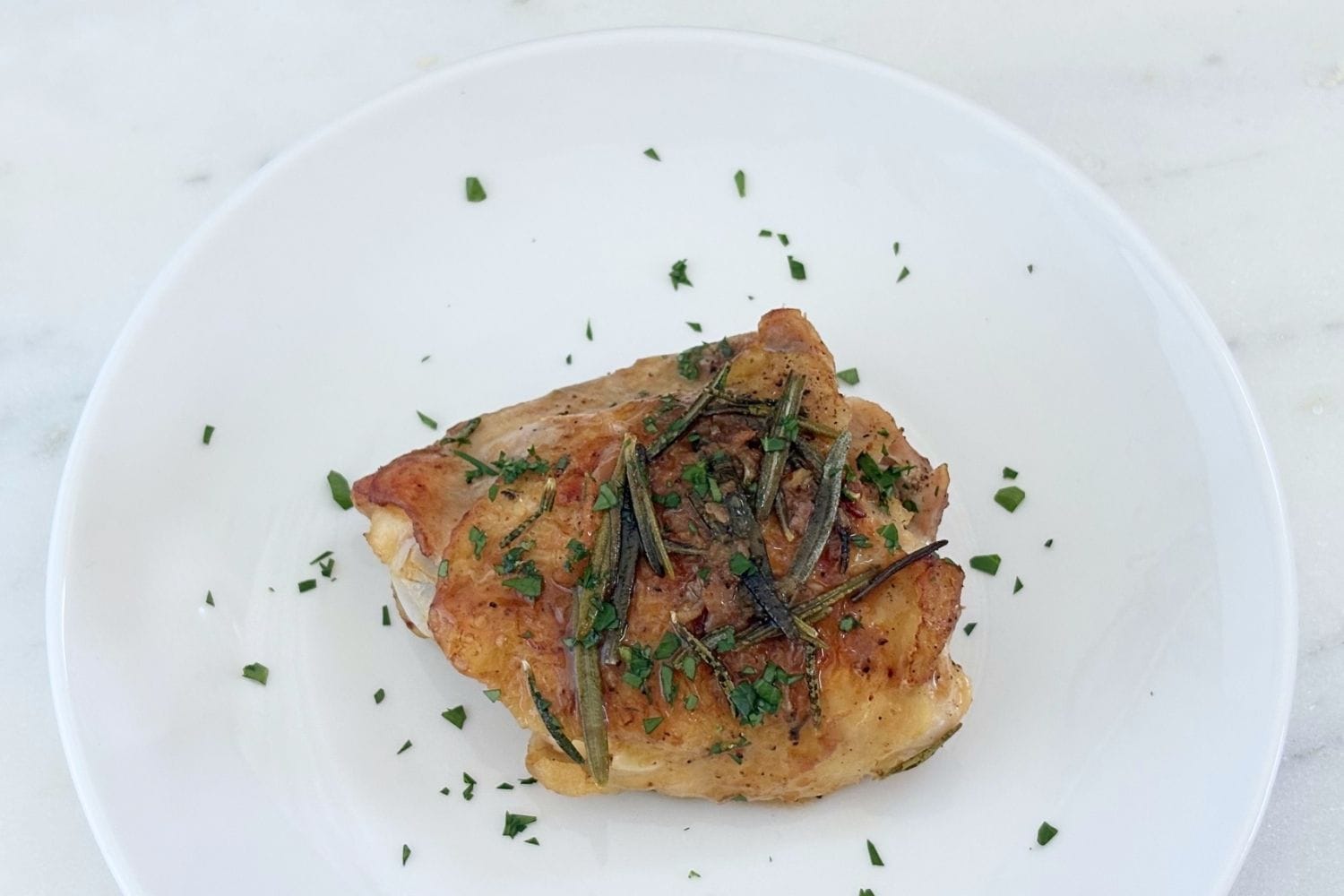 A single bone-in, skin-on chicken thigh served on a white plate with rosemary, pan juices, and chopped parsley.