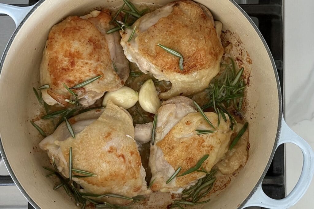 Four browned chicken thighs simmering in an enameled cast iron Dutch oven with garlic cloves, fresh rosemary, and white wine.