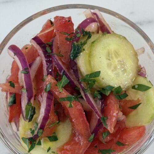 Glass bowl filled with tomato, cucumber, onion, and basil salad made with peeled tomatoes, thin cucumber slices, red onion, and fresh basil.