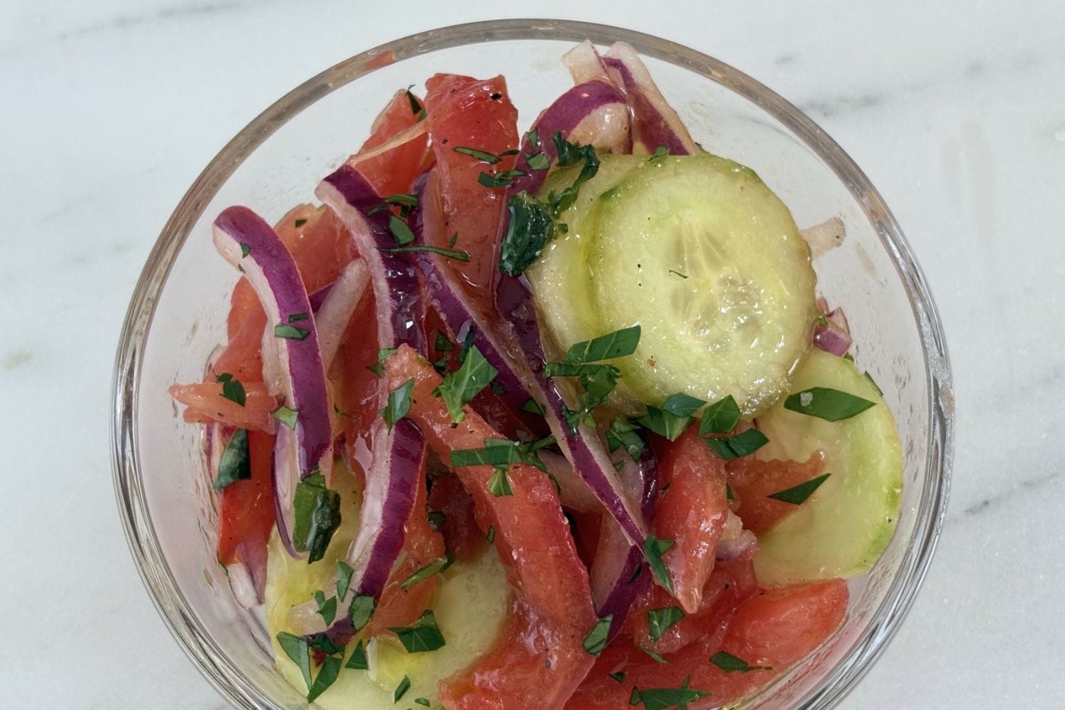 Glass bowl filled with tomato, cucumber, onion, and basil salad made with peeled tomatoes, thin cucumber slices, red onion, and fresh basil.