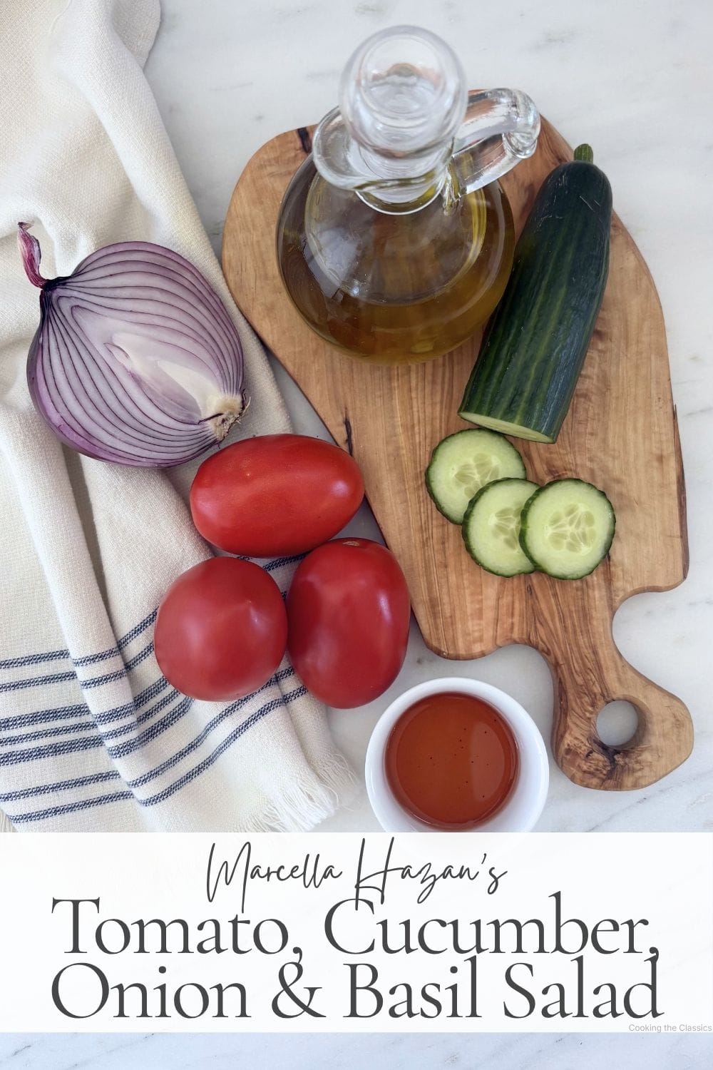 Flatlay of ingredients for Marcella Hazan's tomato, cucumber, onion, and basil salad, including tomatoes, cucumber, red onion, olive oil, vinegar, and a kitchen towel.