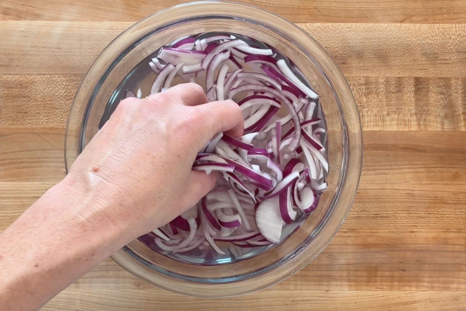 Hand gently squeezing thinly sliced red onion in cold water, a technique used in Marcella Hazan's tomato and cucumber salad to soften the onion's sharpness.