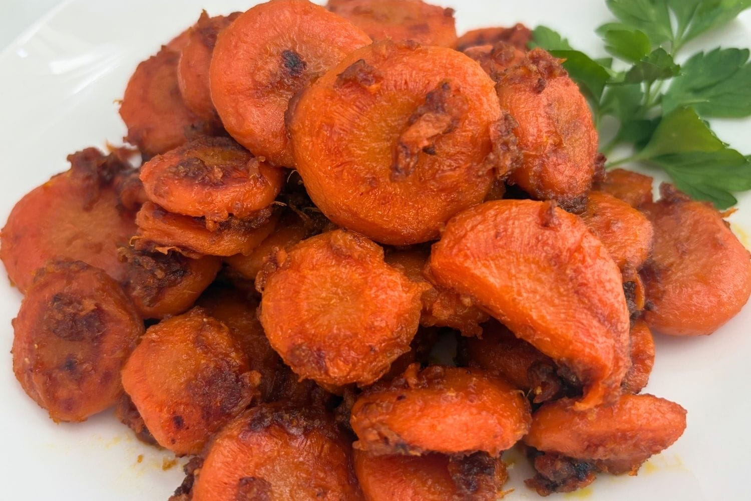 Close-up of braised carrot slices with browned edges and Parmesan cheese, piled on a white plate with fresh parsley.