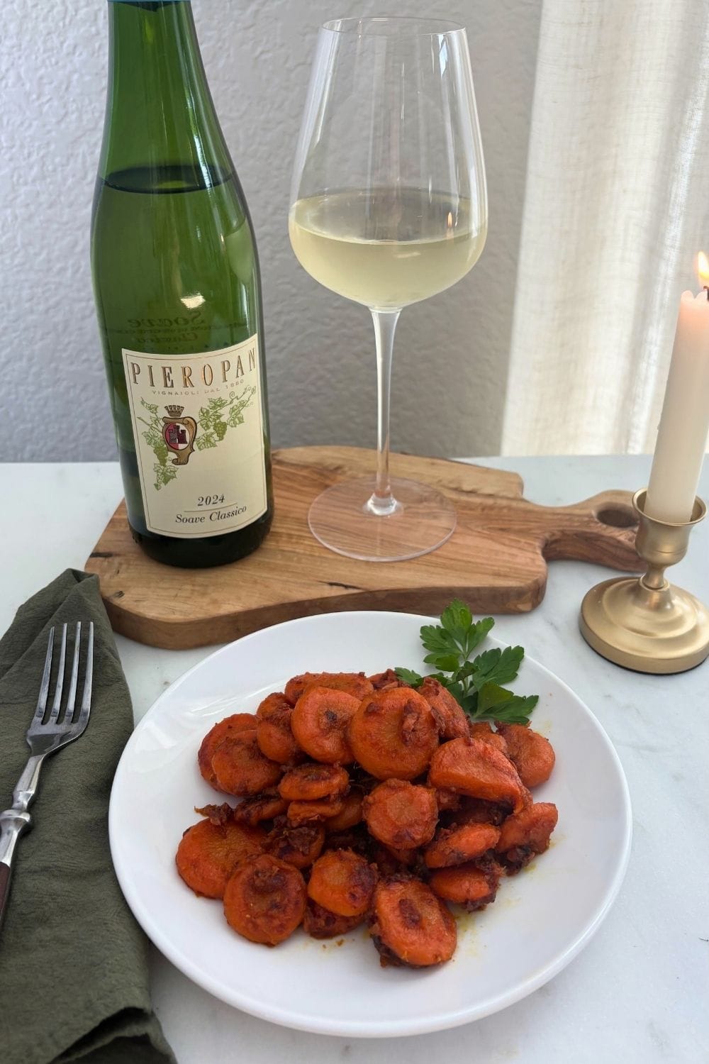 Braised carrots with Parmesan cheese served on a white plate alongside a bottle and glass of Soave, an olive green napkin, and a lit candle.