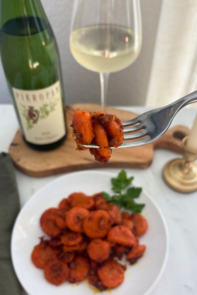 Fork holding a bite of braised carrots with Parmesan cheese above a white plate, with a bottle and glass of Soave softly blurred in the background.