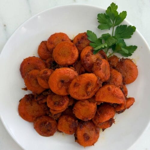 Overhead view of braised carrot slices with browned edges and Parmesan cheese, garnished with fresh parsley on a white plate.