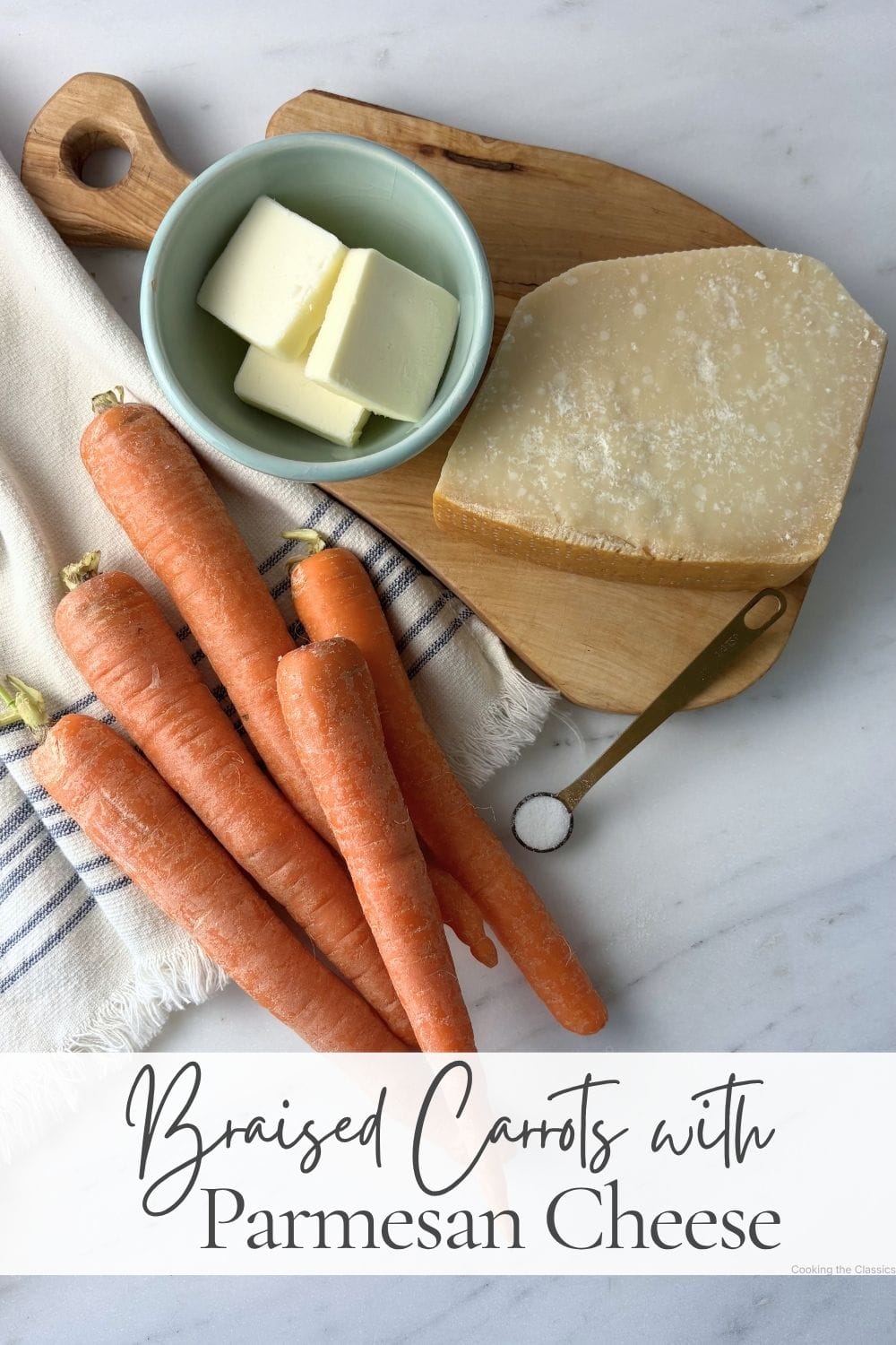 Flat lay of the ingredients for Marcella Hazan's Braised Carrots with Parmesan Cheese, including whole carrots, butter, a wedge of Parmigiano-Reggiano, and salt arranged on an olive wood board over a striped kitchen towel on a marble countertop.