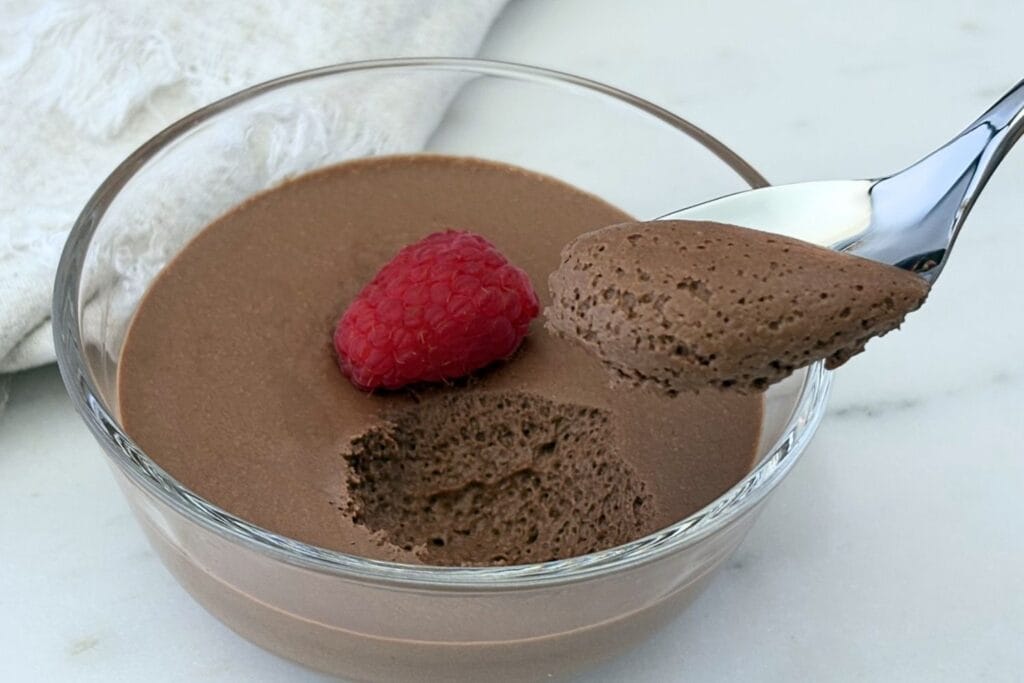 Chocolate mousse in a glass dessert bowl topped with a fresh raspberry, with a spoon lifting a light, airy bite to show the mousse's smooth texture.