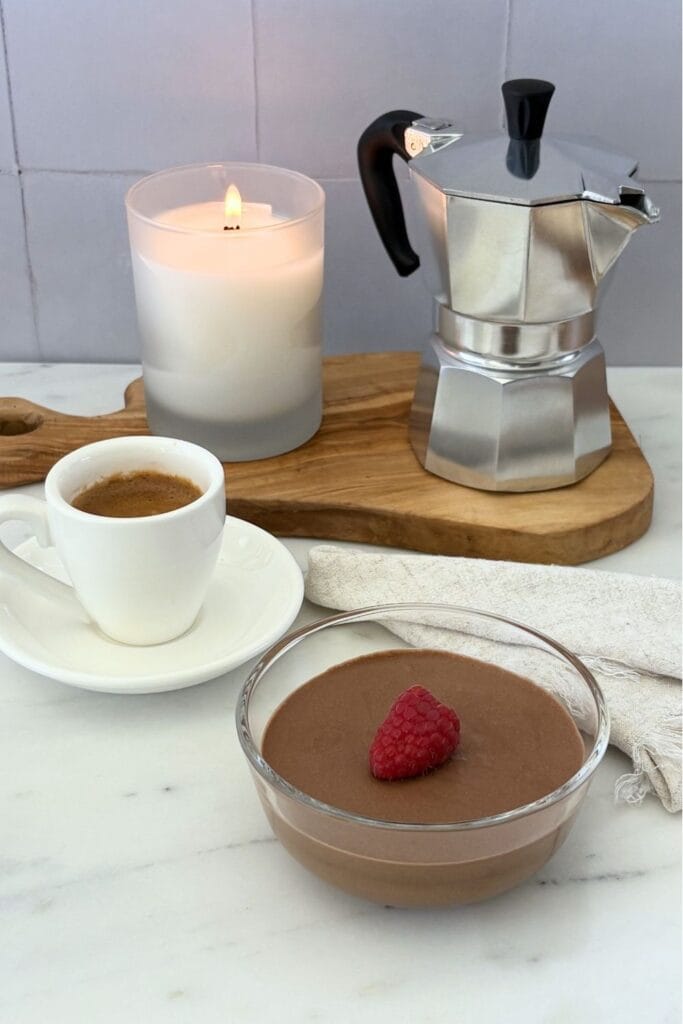 Italian chocolate mousse topped with a fresh raspberry served alongside a cup of espresso and a stovetop moka pot on an olive wood board.