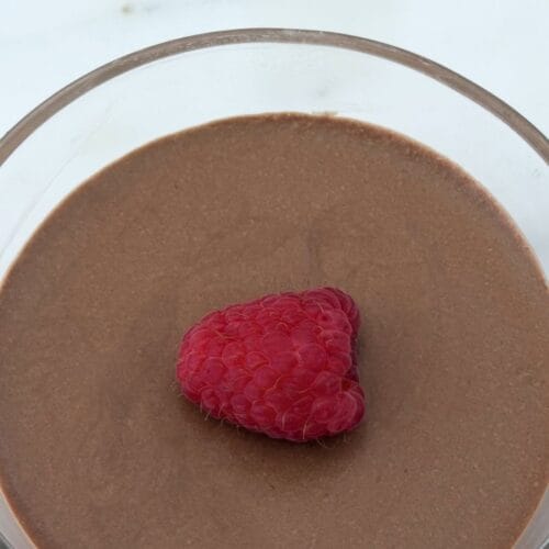 Overhead view of Italian chocolate mousse in a glass dessert bowl garnished with a single fresh raspberry on a smooth chocolate surface.