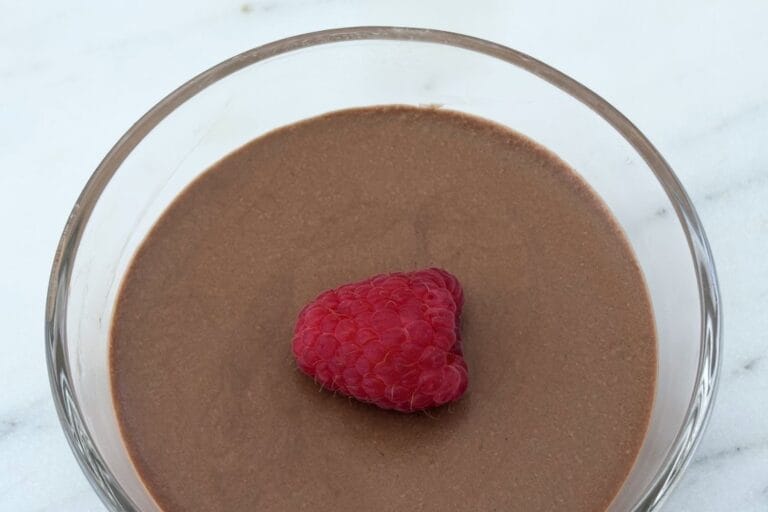 Overhead view of Italian chocolate mousse in a glass dessert bowl garnished with a single fresh raspberry on a smooth chocolate surface.