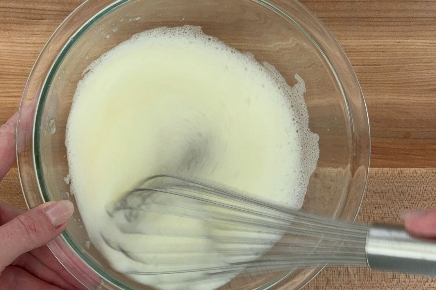 Hands whisking beaten egg whites in a clear glass bowl while preparing Marcella Hazan's Italian chocolate mousse.