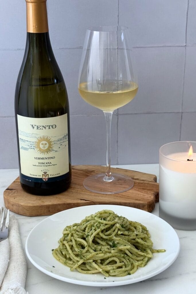 Plate of basil pesto spaghetti served with a glass and bottle of Italian Vermentino wine on a wooden board beside a lit candle.