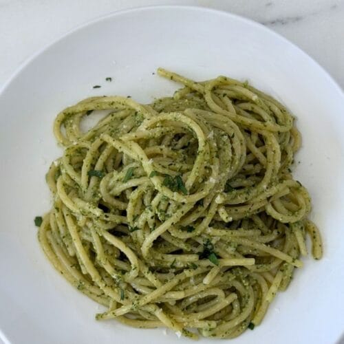 Overhead view of spaghetti tossed with homemade basil pesto in a white pasta bowl, finished with grated Parmigiano-Reggiano and chopped parsley.