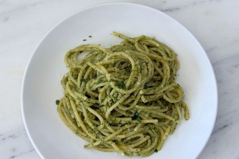 Overhead view of spaghetti tossed with homemade basil pesto in a white pasta bowl, finished with grated Parmigiano-Reggiano and chopped parsley.