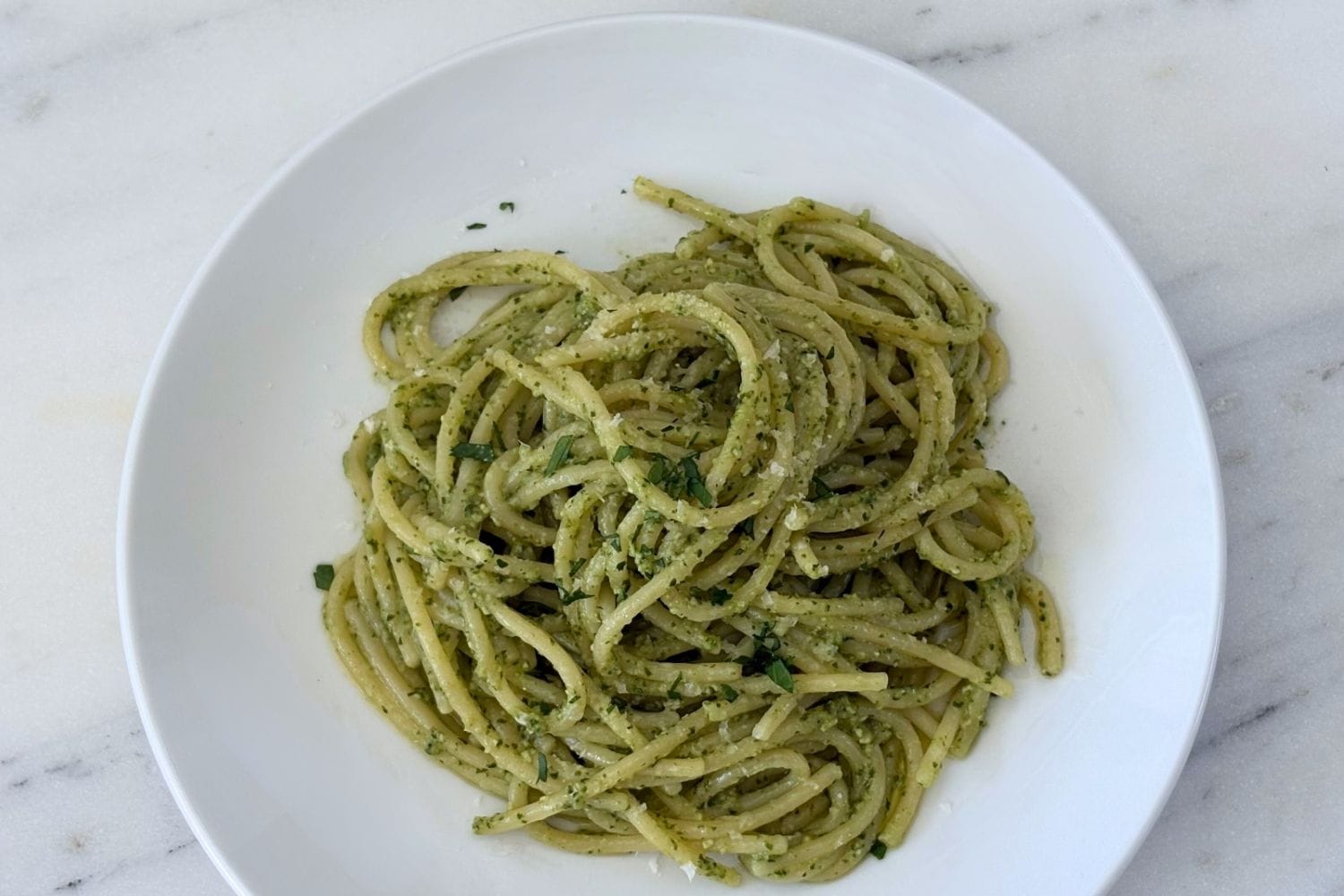Overhead view of spaghetti tossed with homemade basil pesto in a white pasta bowl, finished with grated Parmigiano-Reggiano and chopped parsley.