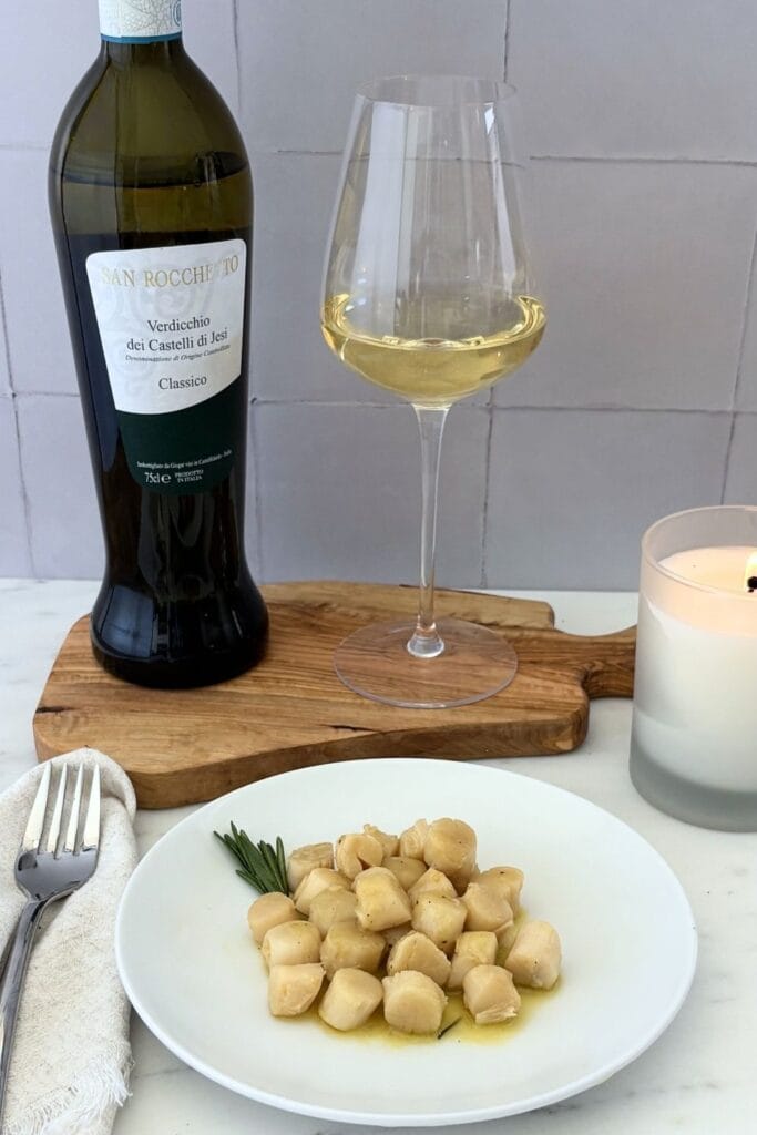 Plate of sautéed scallops with rosemary and lemon beside a bottle and glass of Verdicchio wine.