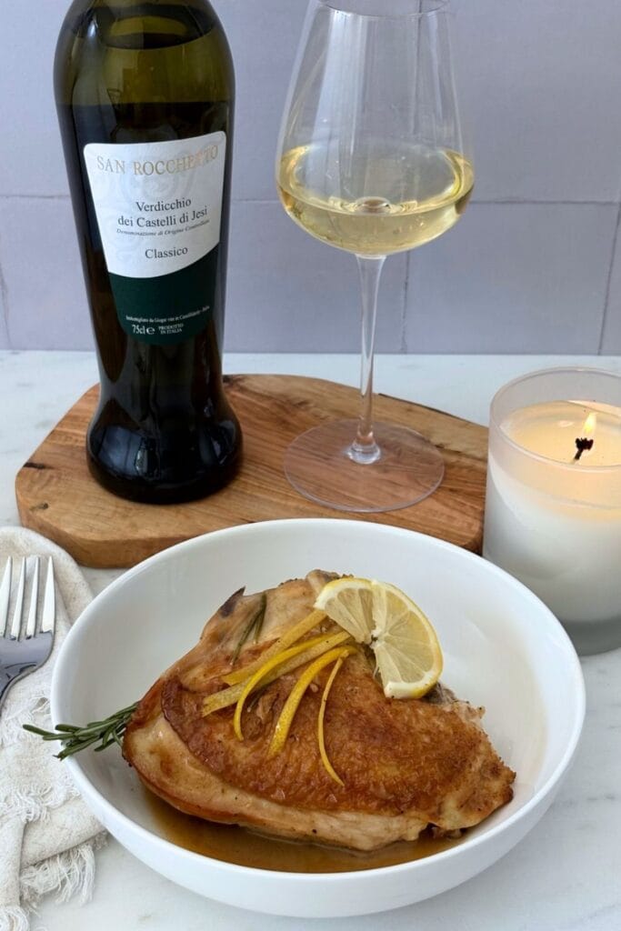 Golden-brown fricasseed chicken with rosemary and lemon served in a white bowl with pan juices, lemon slices, and a glass of Verdicchio.