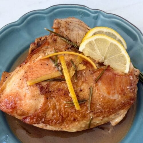 Golden roasted chicken with rosemary, lemon zest, and lemon slices served on a blue plate with pan sauce.