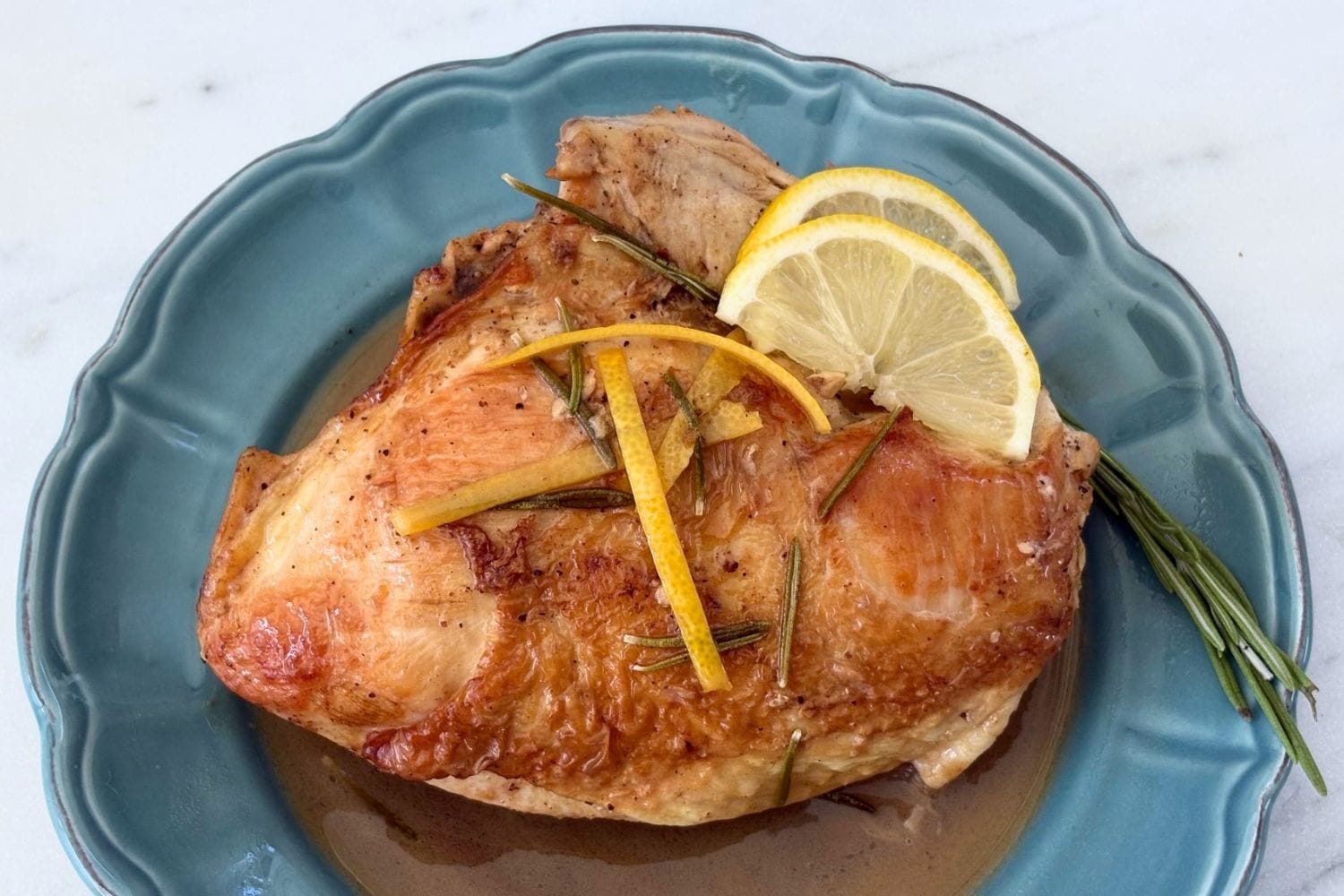 Golden roasted chicken with rosemary, lemon zest, and lemon slices served on a blue plate with pan sauce.