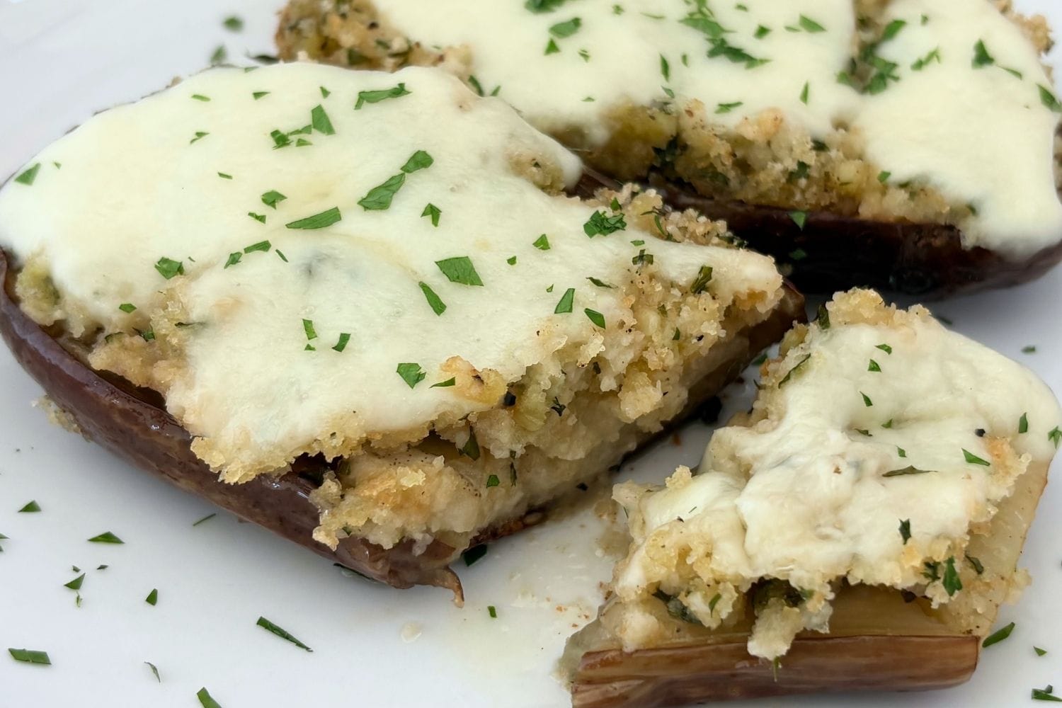 Sautéed baby eggplant halves topped with seasoned breadcrumbs, melted mozzarella, and chopped parsley on a white plate.