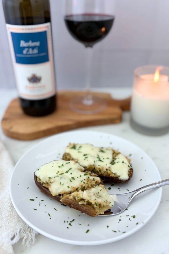 Sautéed baby eggplant halves topped with breadcrumbs, melted mozzarella, and parsley, plated beside a glass and bottle of Barbera.