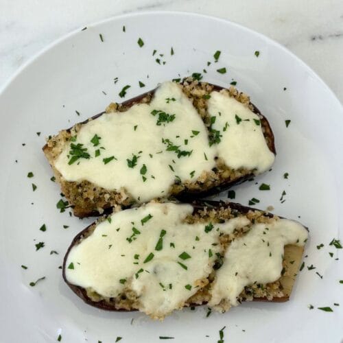 Sautéed baby eggplant halves topped with seasoned breadcrumbs, melted mozzarella, and chopped parsley on a white plate.