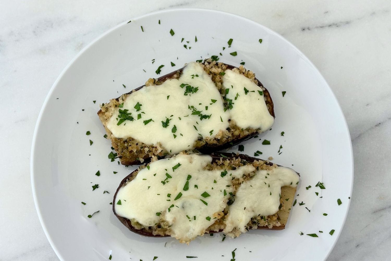Sautéed baby eggplant halves topped with seasoned breadcrumbs, melted mozzarella, and chopped parsley on a white plate.