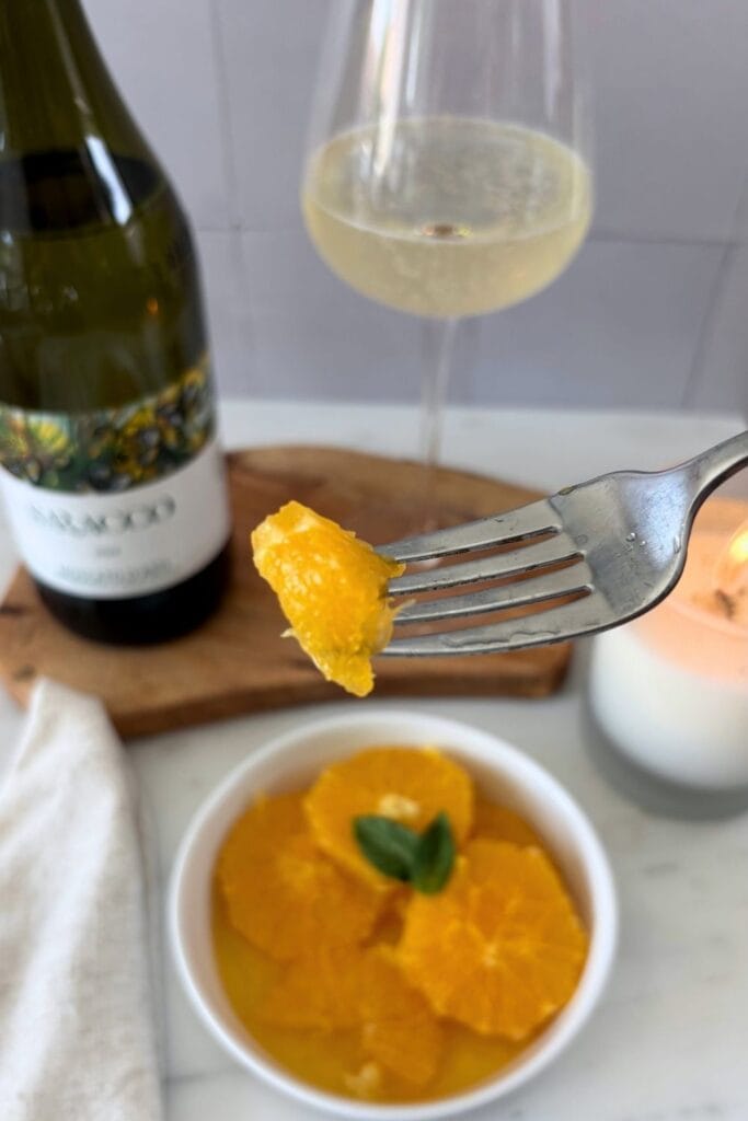 A fork holding a macerated orange slice above a bowl of oranges, with Moscato d’Asti and a wine glass in the background.