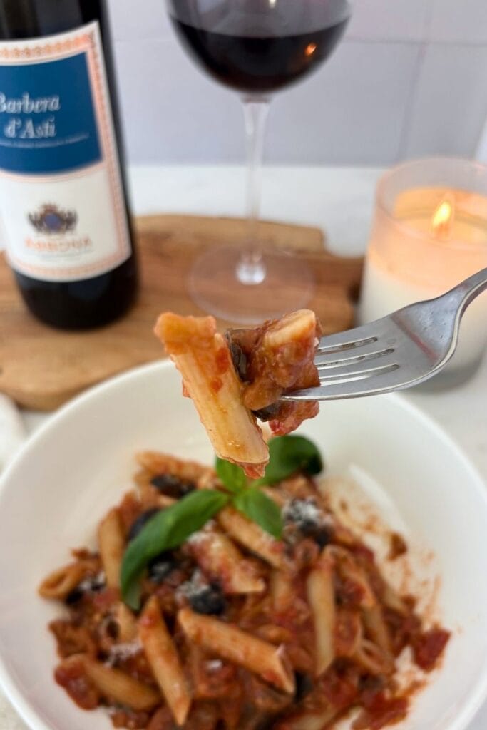 Fork holding a bite of penne with eggplant and tomato sauce above a bowl of pasta, with Barbera wine and a candle in the background.