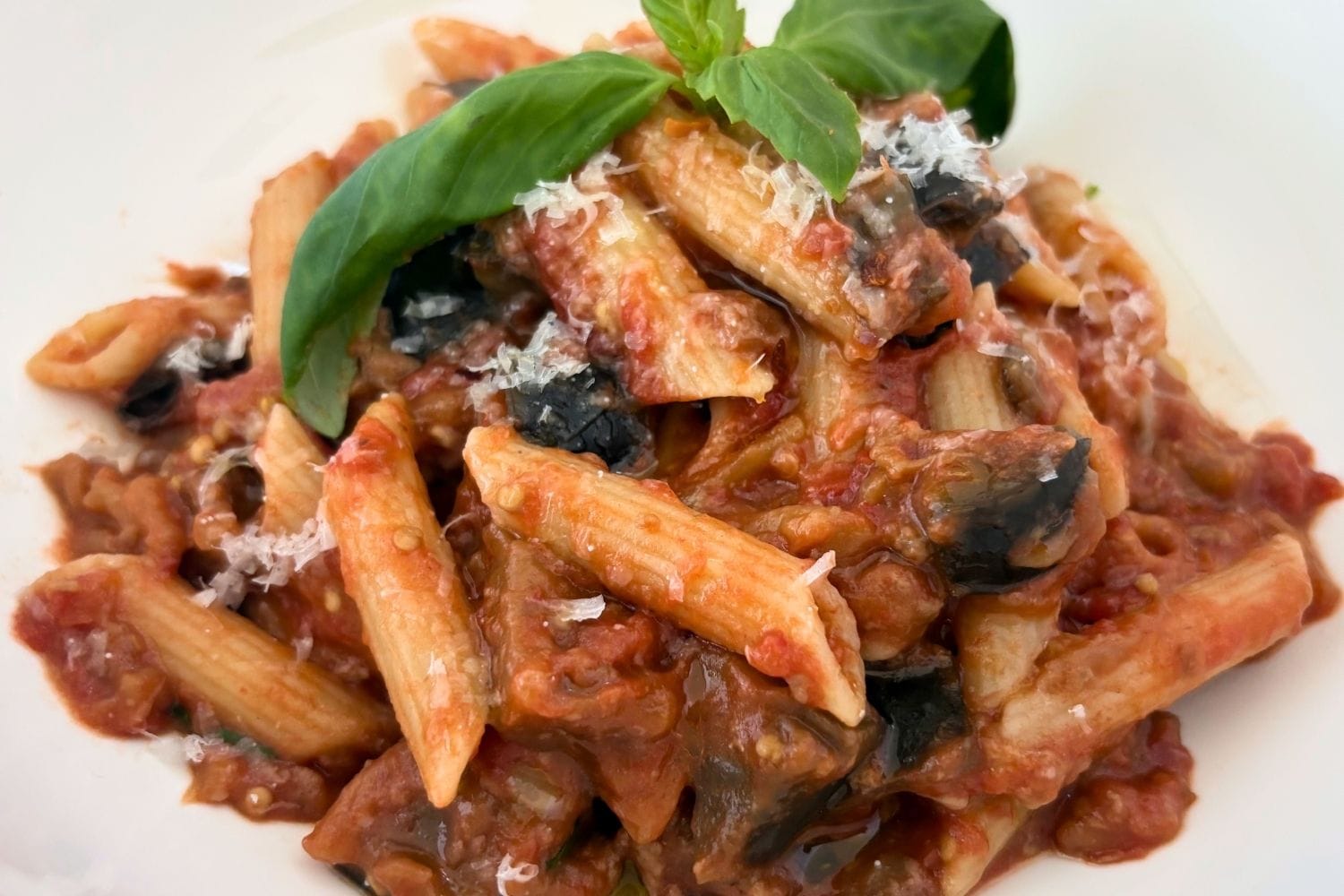 Bowl of penne coated in tomato and eggplant sauce, topped with grated Pecorino Romano and fresh basil.