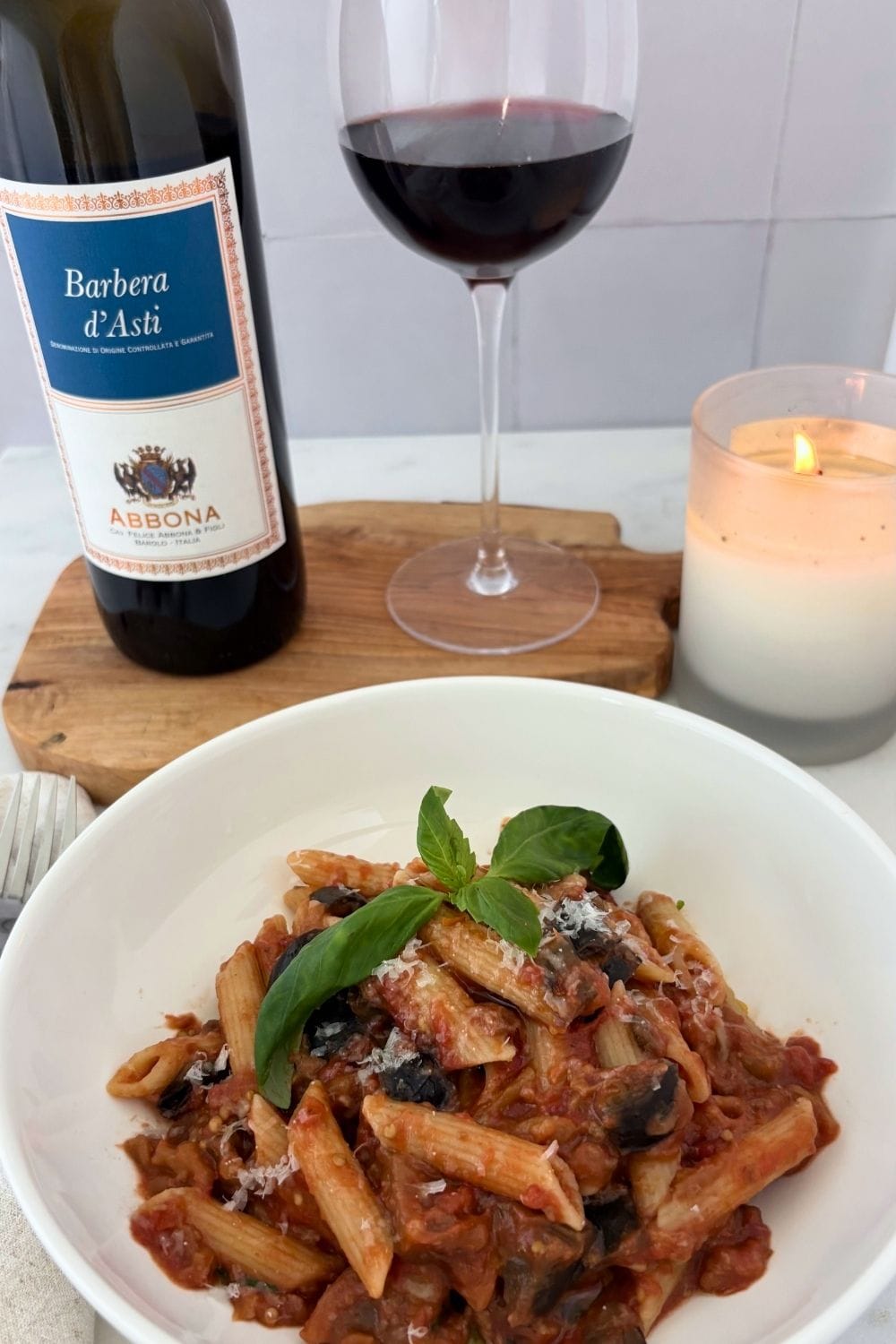 Bowl of eggplant, tomato, and mozzarella pasta with fresh basil, served alongside a bottle and glass of Barbera and a lit candle.