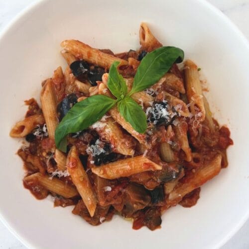 Overhead view of penne with chunky eggplant and tomato sauce, grated Pecorino Romano, and fresh basil in a white bowl.