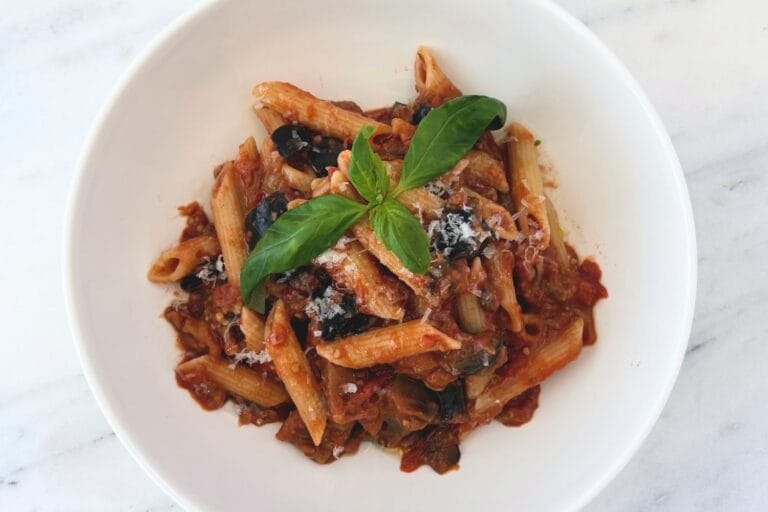 Overhead view of penne with chunky eggplant and tomato sauce, grated Pecorino Romano, and fresh basil in a white bowl.
