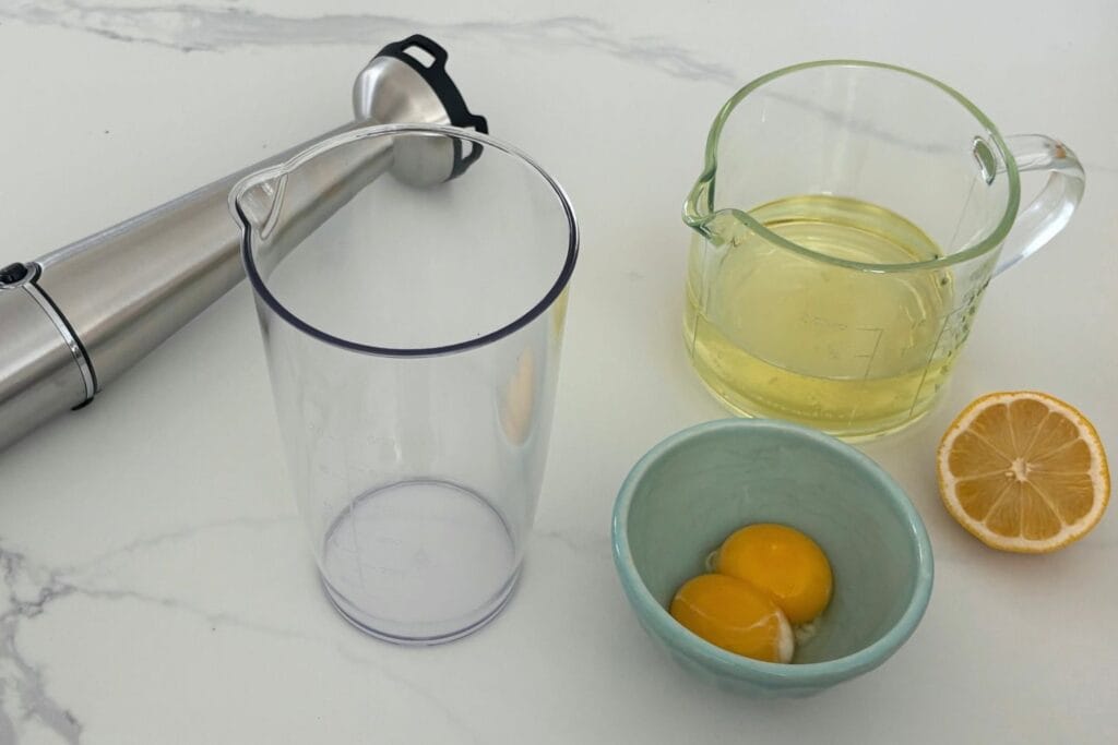 Egg yolks, vegetable oil, lemon, immersion blender, and blending container for making homemade mayonnaise.