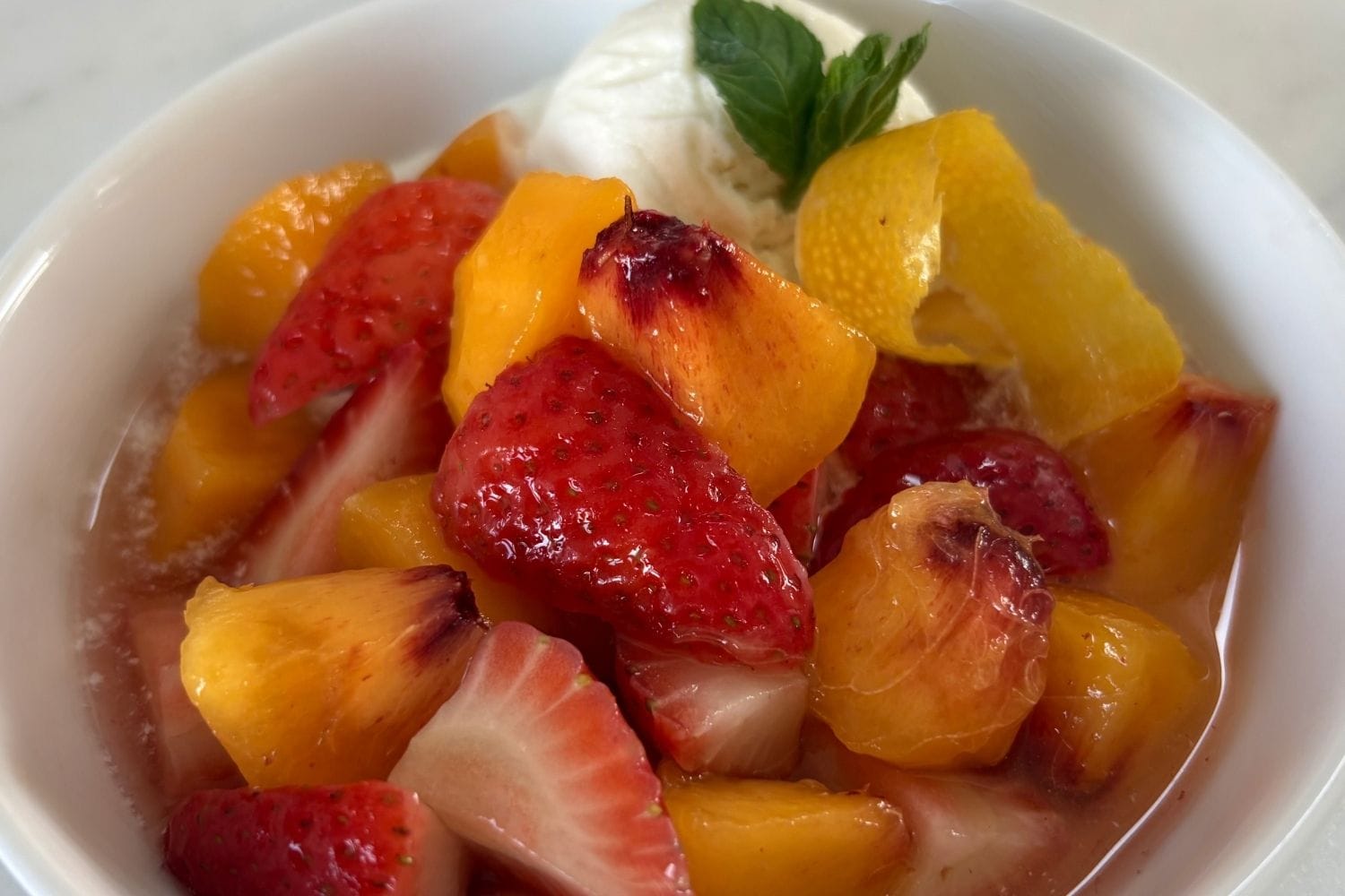 Close-up of peaches and strawberries in sweet white wine, served with vanilla ice cream, mint, and lemon peel.