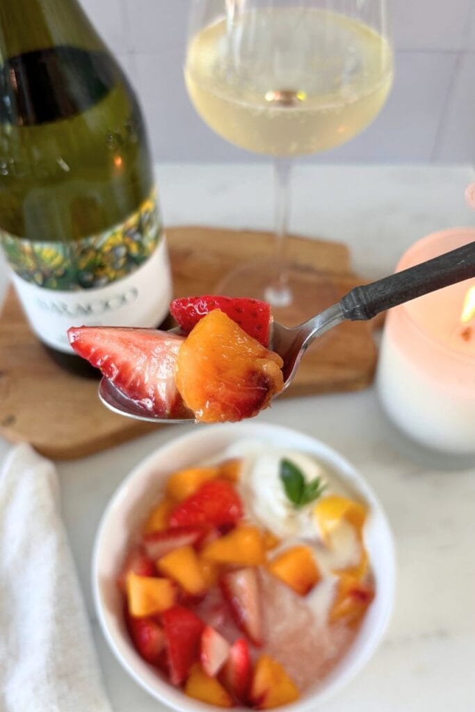Spoonful of macerated peaches and strawberries held above a bowl of fruit, with Moscato d’Asti and a glass of wine in the background.