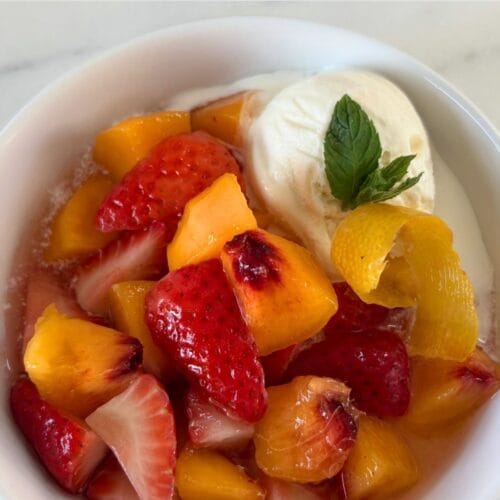 Overhead view of peaches and strawberries in sweet white wine with vanilla ice cream, fresh mint, and a curl of lemon peel.