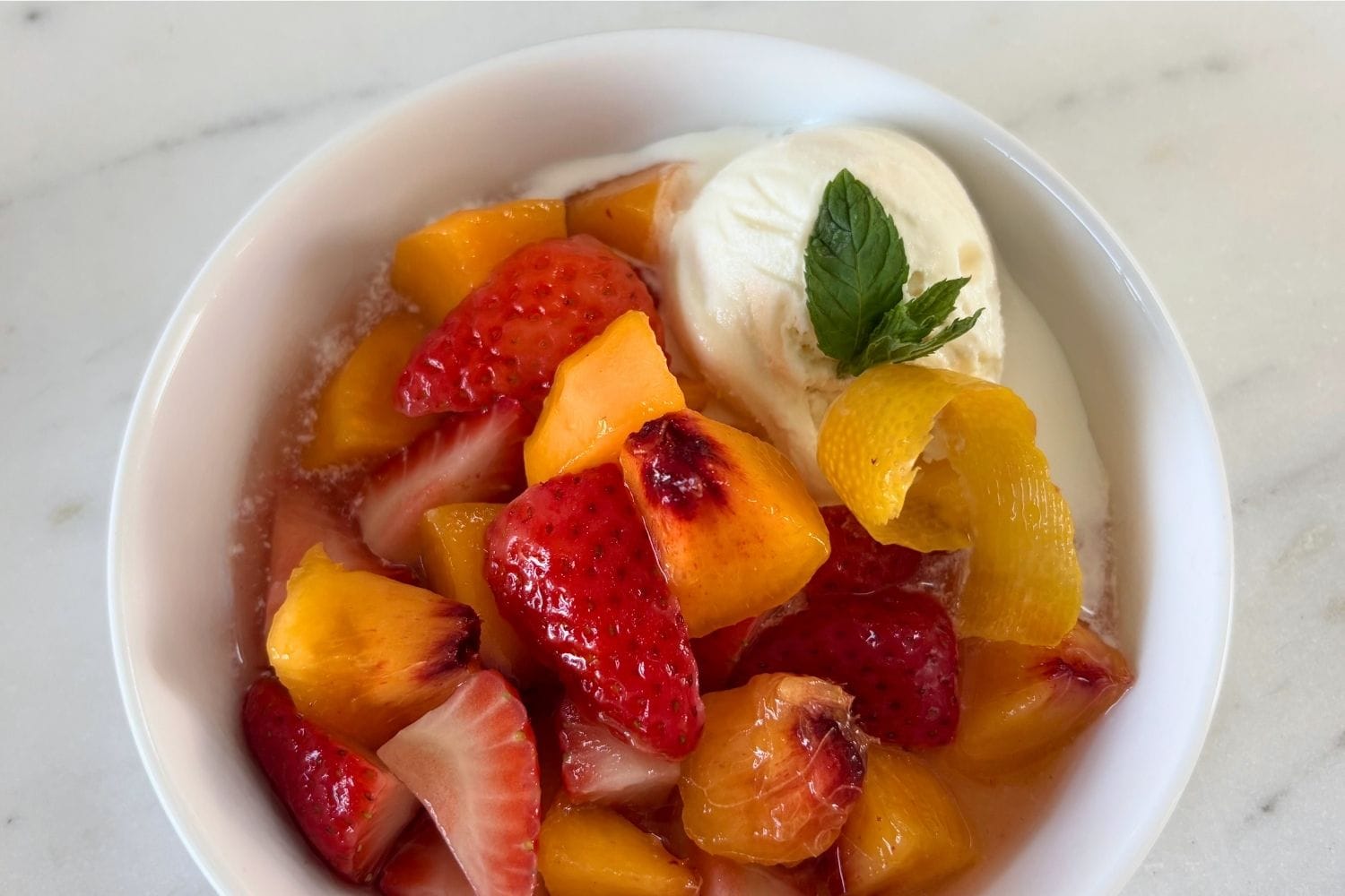 Overhead view of peaches and strawberries in sweet white wine with vanilla ice cream, fresh mint, and a curl of lemon peel.
