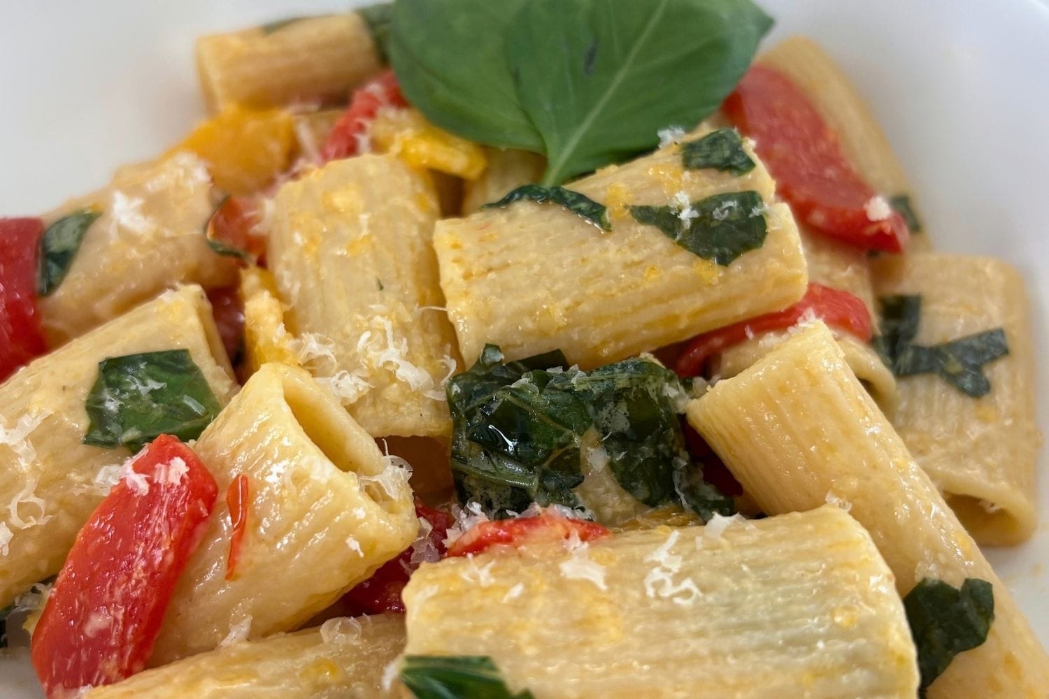 Close-up of rigatoni with red and yellow bell peppers, torn basil leaves, and freshly grated Parmesan cheese.