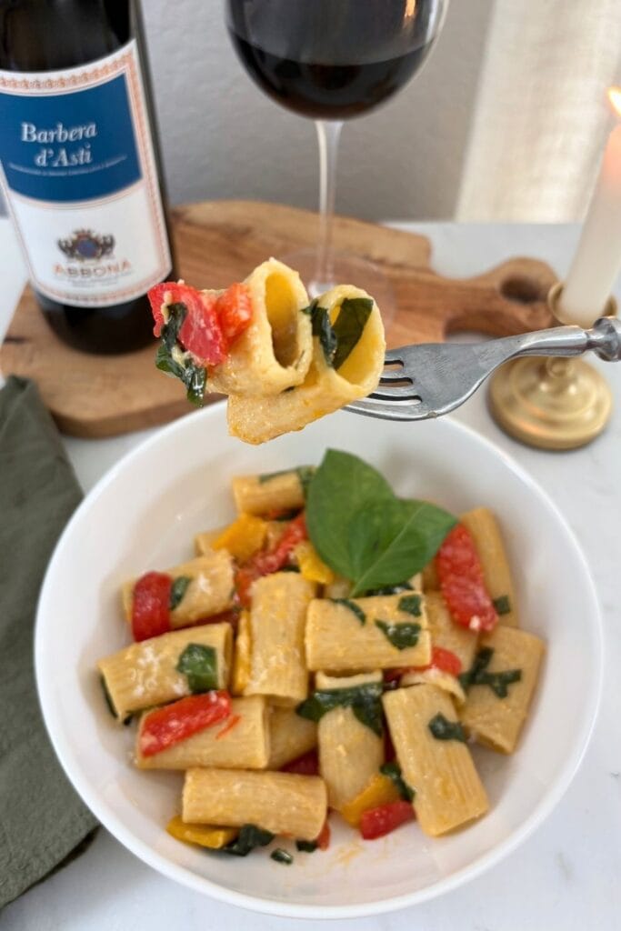 Fork holding rigatoni with red bell pepper and fresh basil above a bowl of pasta, with a glass of Barbera, wine bottle, and lit candle in the background.