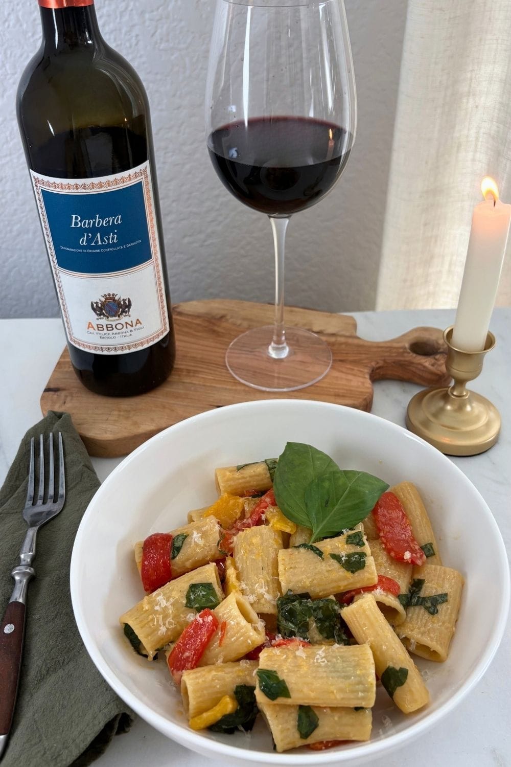 White bowl of rigatoni with red and yellow bell peppers, fresh basil, and grated Parmesan, served alongside a bottle and glass of Barbera wine and a lit candle.