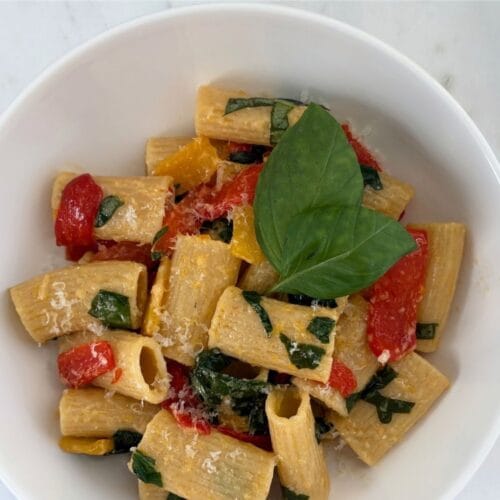Overhead view of rigatoni tossed with red and yellow bell peppers, fresh basil, and Parmesan cheese in a white bowl on a marble surface.