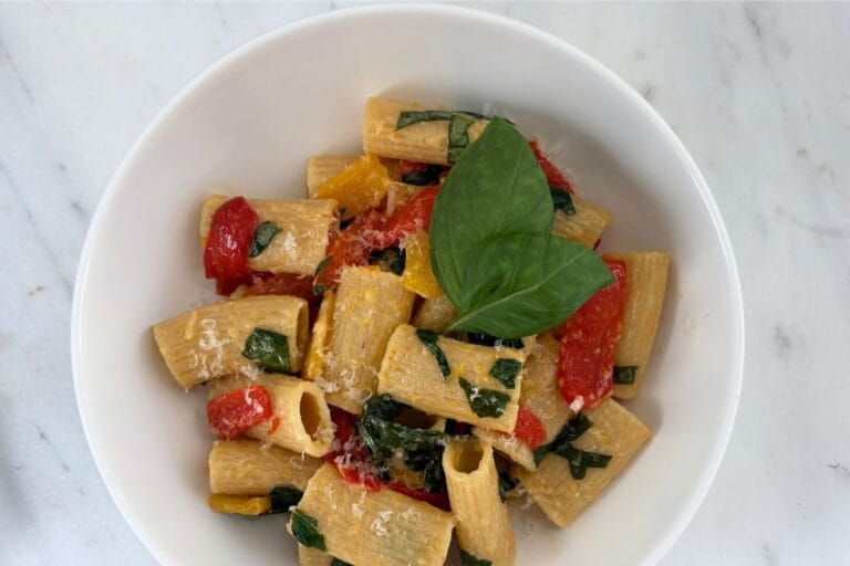 Overhead view of rigatoni tossed with red and yellow bell peppers, fresh basil, and Parmesan cheese in a white bowl on a marble surface.