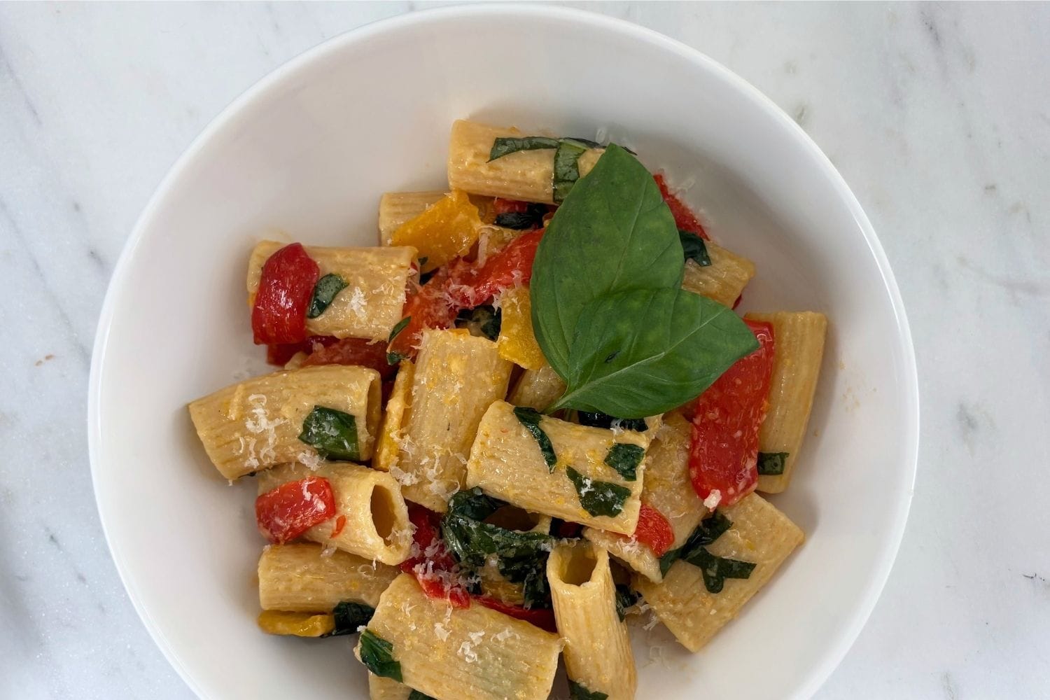Overhead view of rigatoni tossed with red and yellow bell peppers, fresh basil, and Parmesan cheese in a white bowl on a marble surface.