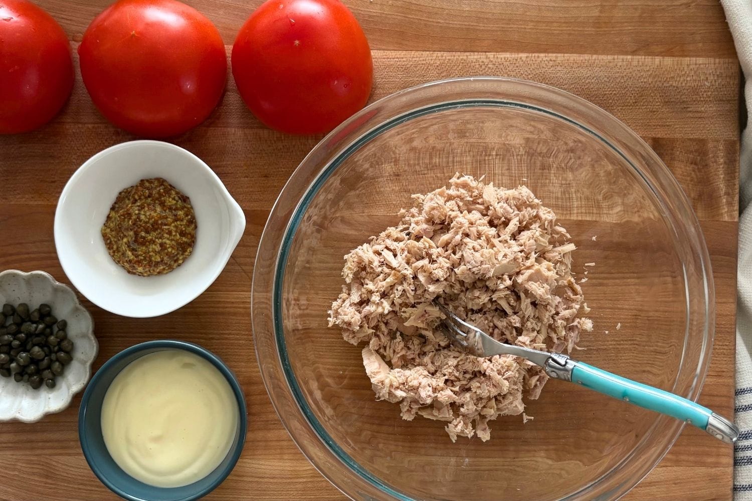 Flaked tuna in a mixing bowl with homemade mayonnaise, capers, mustard, and tomatoes arranged nearby.